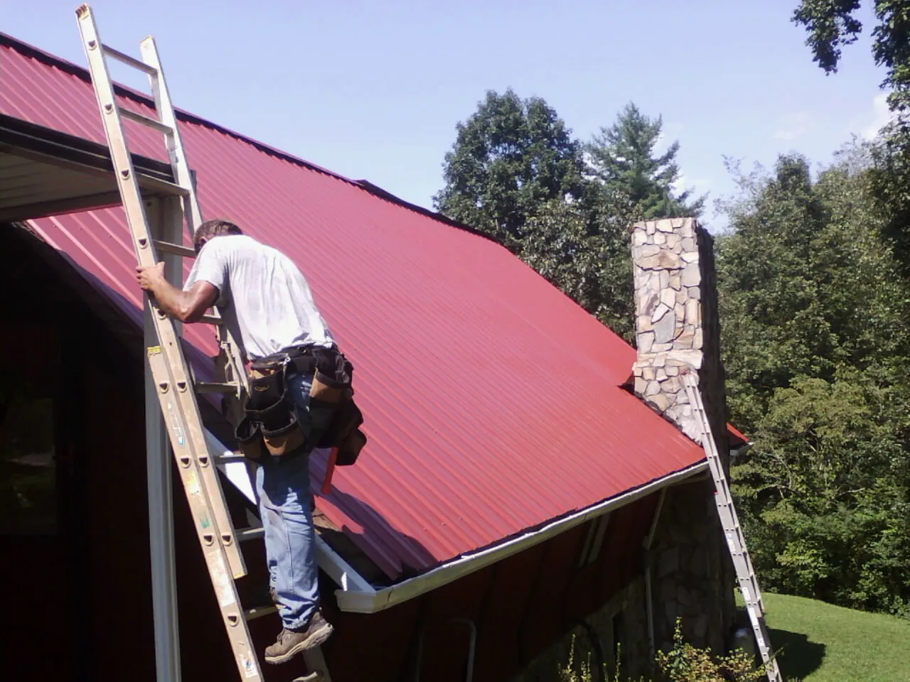 Roofer on a ladder beside a red metal roof and stone chimney — Soffit & Fascia Repair by Roofing Brothers in Old Station